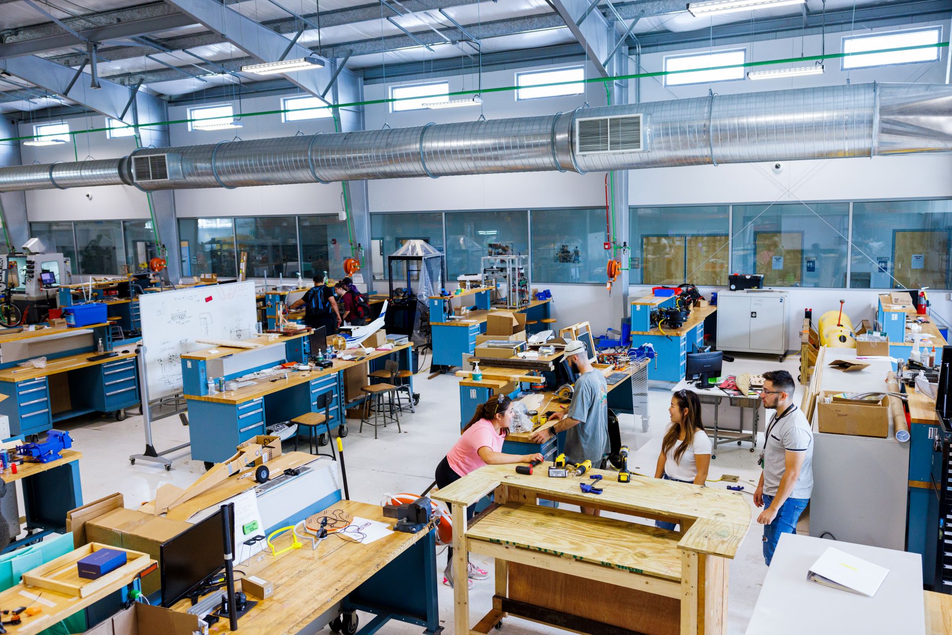 Busy engineering workshop with students working at various stations, surrounded by tools and projects under bright industrial lights